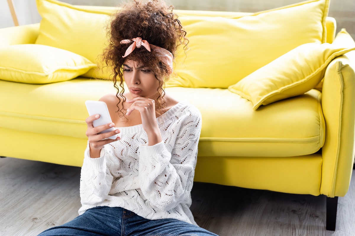 worried woman looking at smartphone near yellow sofa
