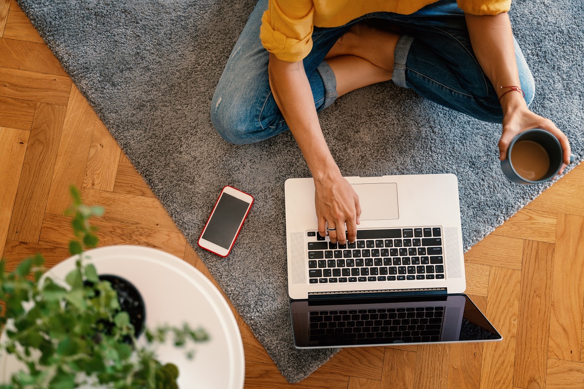 woman in a yellow shirt works on a laptop with a mug of coffee at home in the living room on the carpet