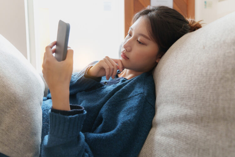 Young Asian woman serious using phone while lying on sofa.
