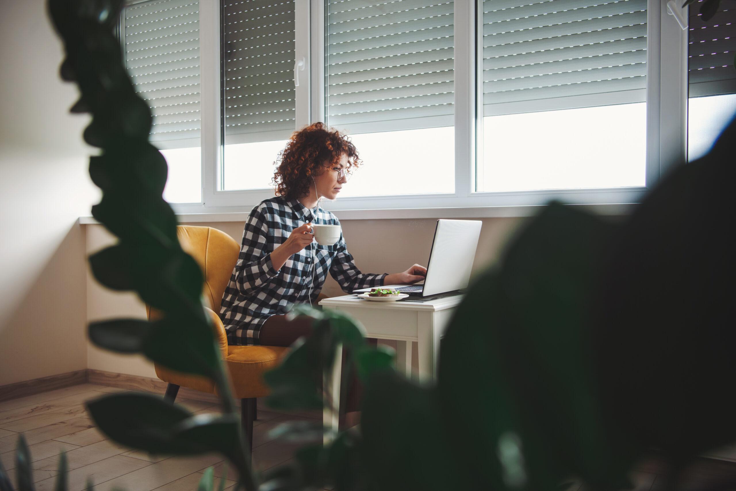 Caucasian woman in eyewear working on laptop while sitting on armchair near window