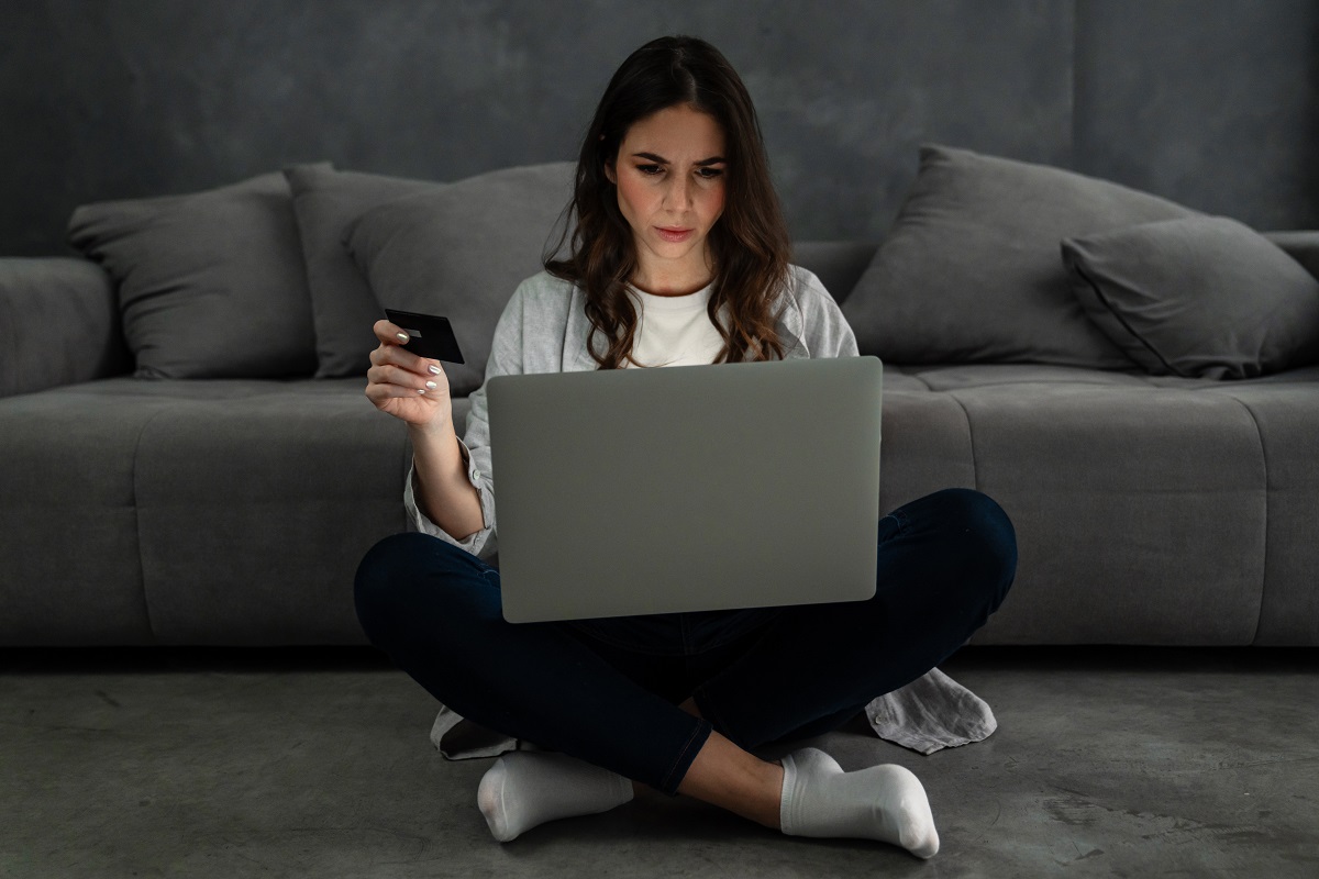 Upset confused woman sitting on the floor, laptop on her lap, and credit card on one hand