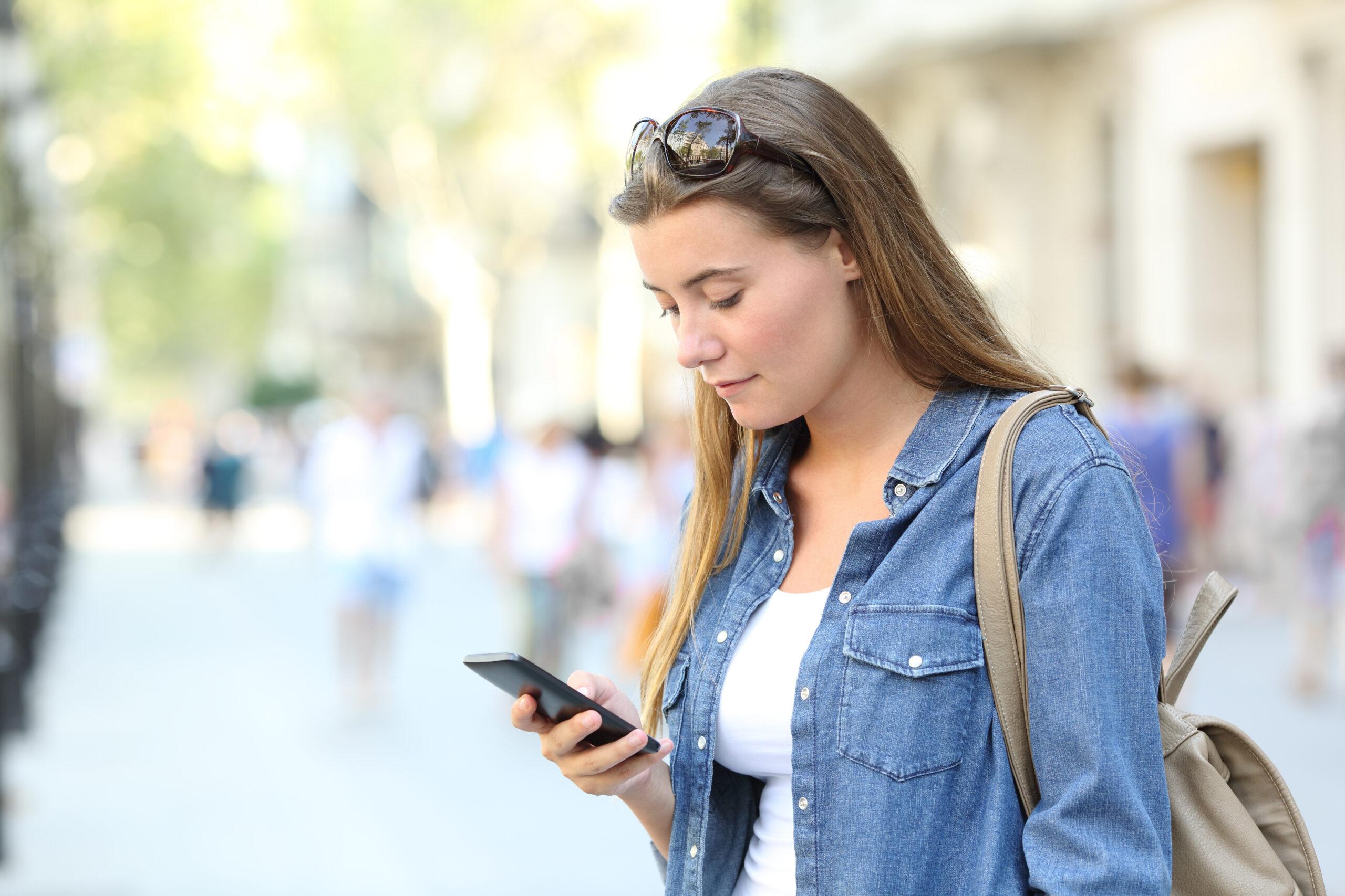 Chica revisando el contenido de un teléfono inteligente en una calle de la ciudad