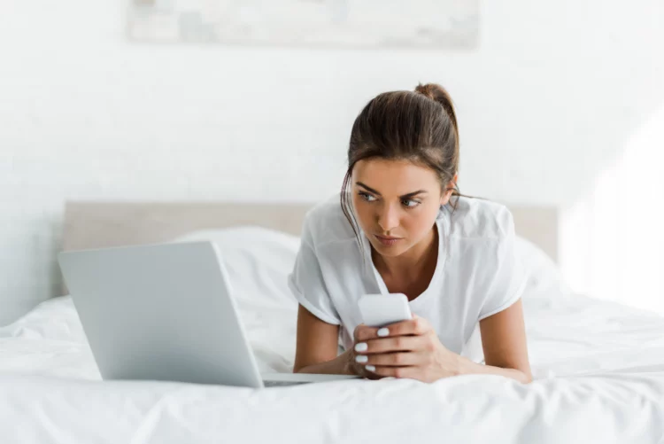 beautiful serious girl using smartphone and laptop in bed 
