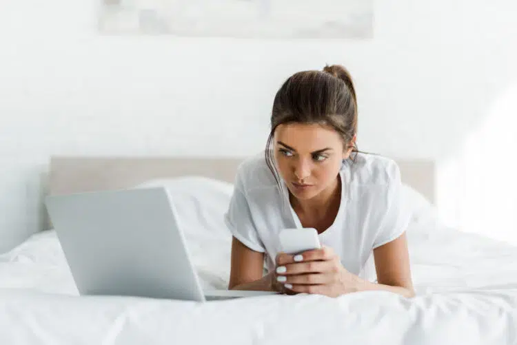 beautiful serious girl using smartphone and laptop in bed