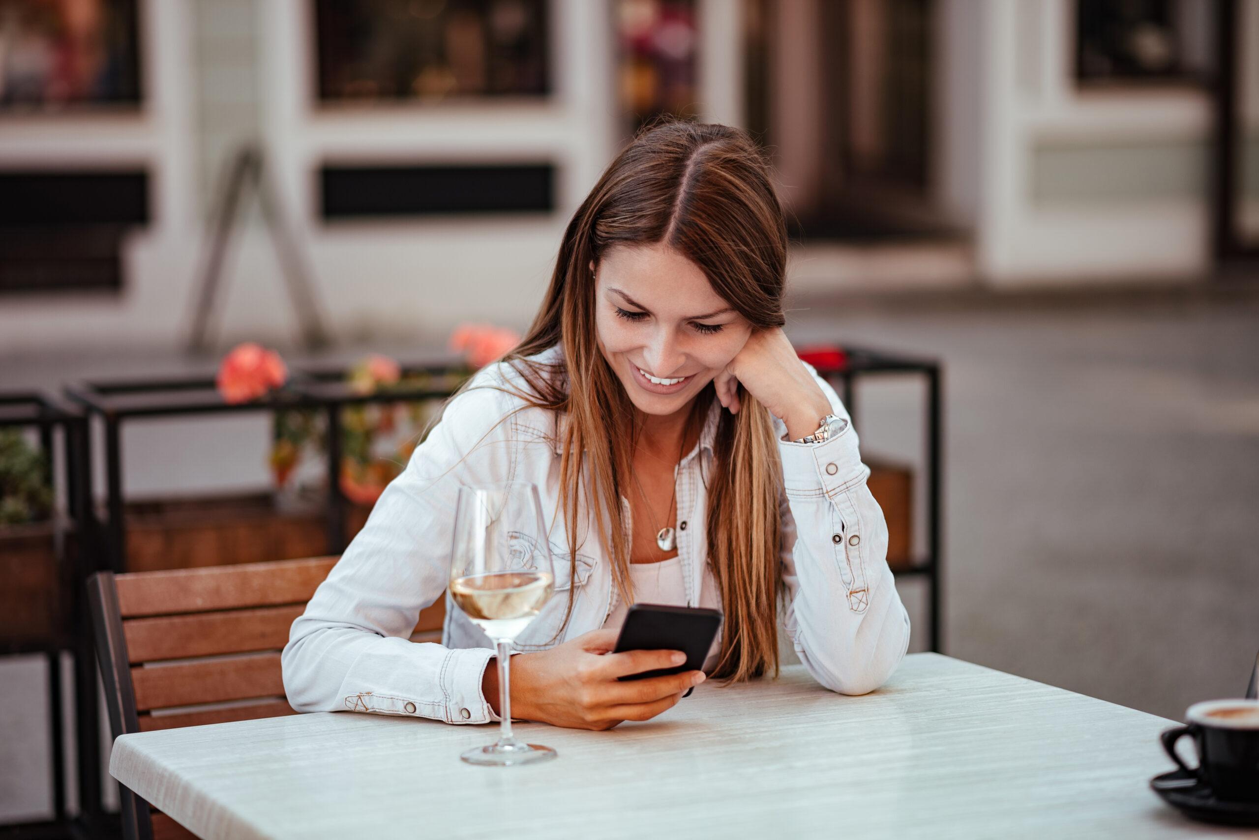 Happy young woman sitting in the restaurant outdoors, looking at smartphone. Glass of white wine on the table.
