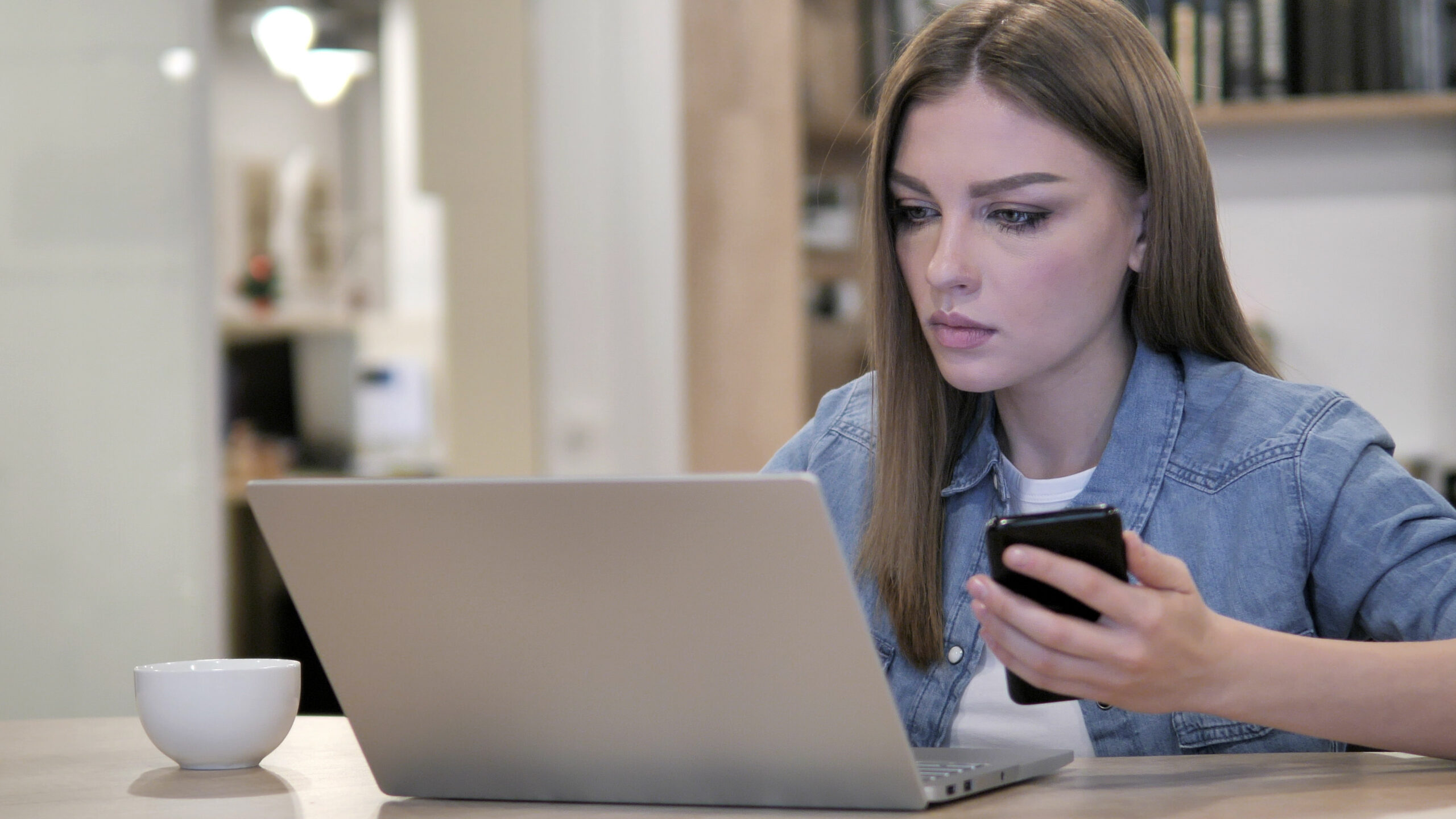 young woman with a curious look using laptop and smartphone at a desk