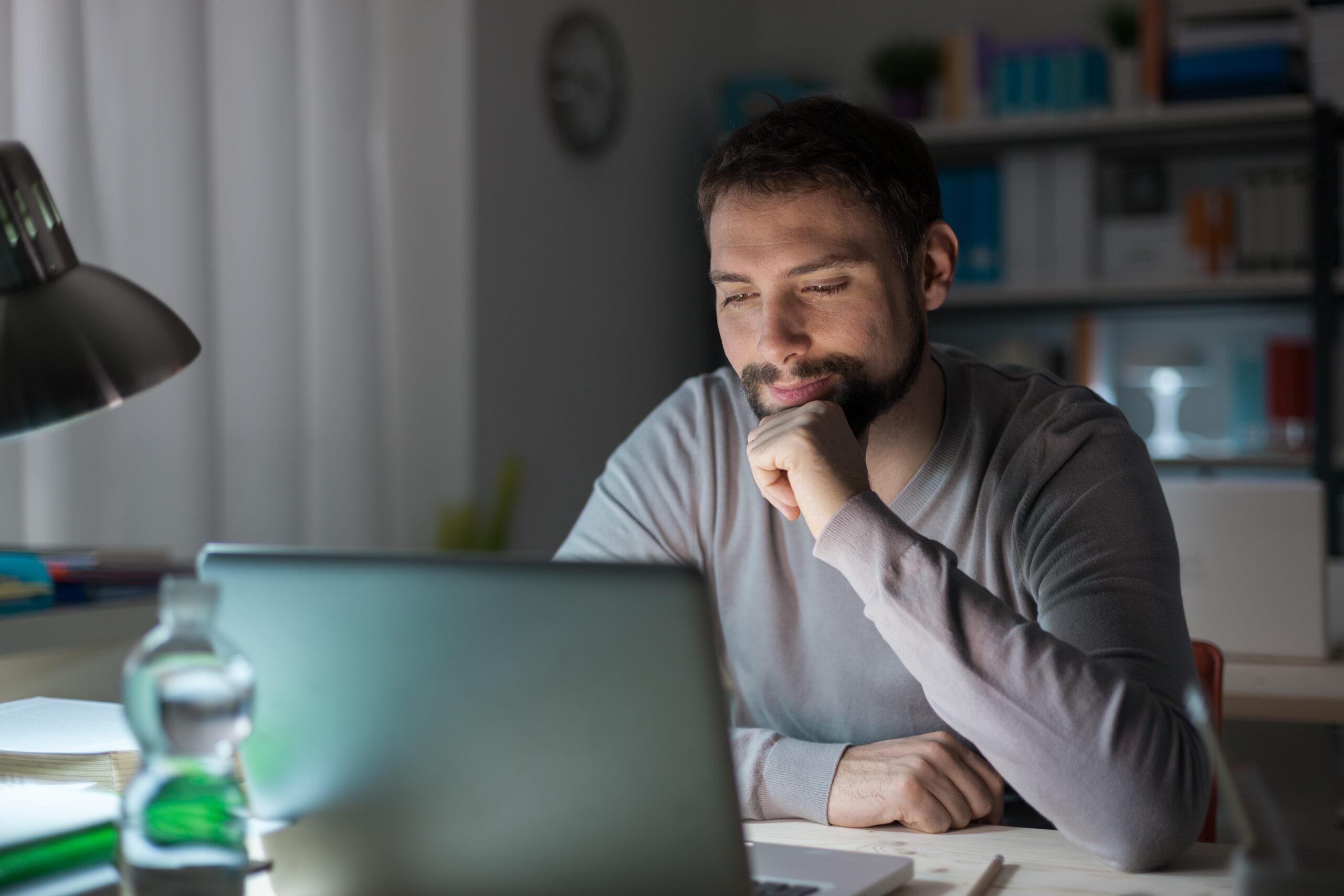 Man with left hand on chin watching a video on his laptop late at night