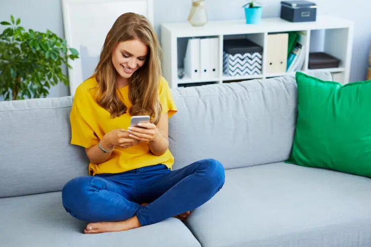 beautiful young woman looking at phone and smiling while sitting on couch at home