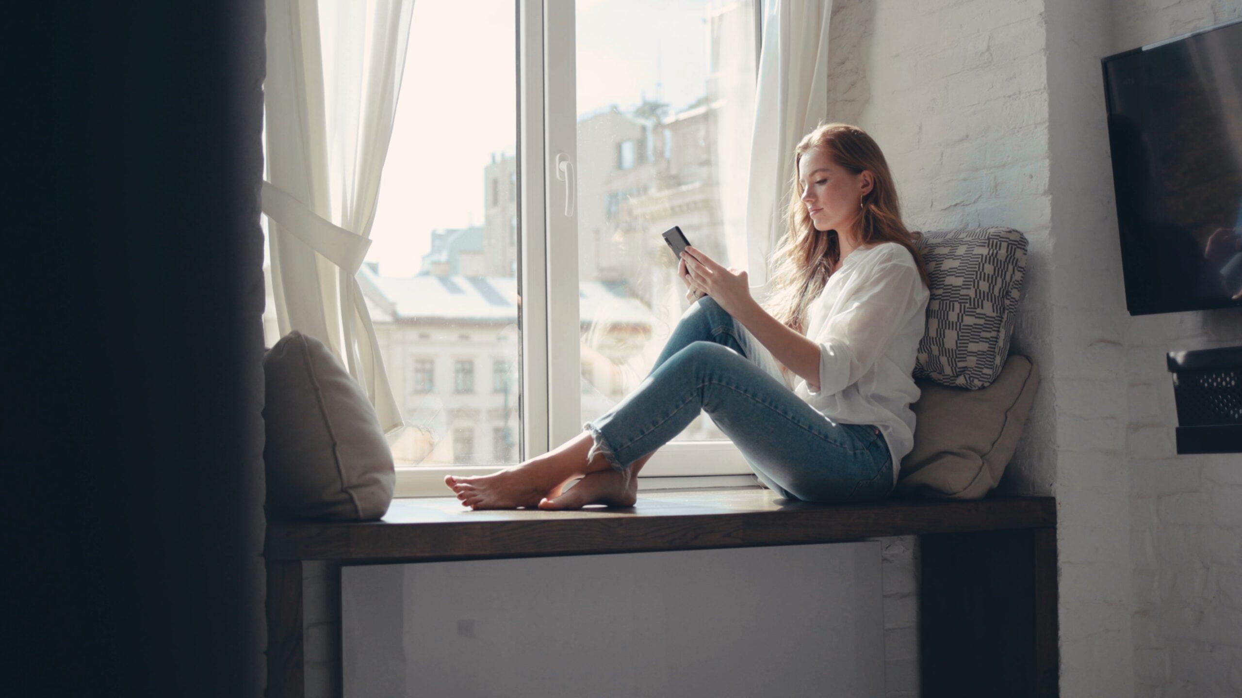 Cheerful young woman sitting on a windowsill at home and texting on her phone 