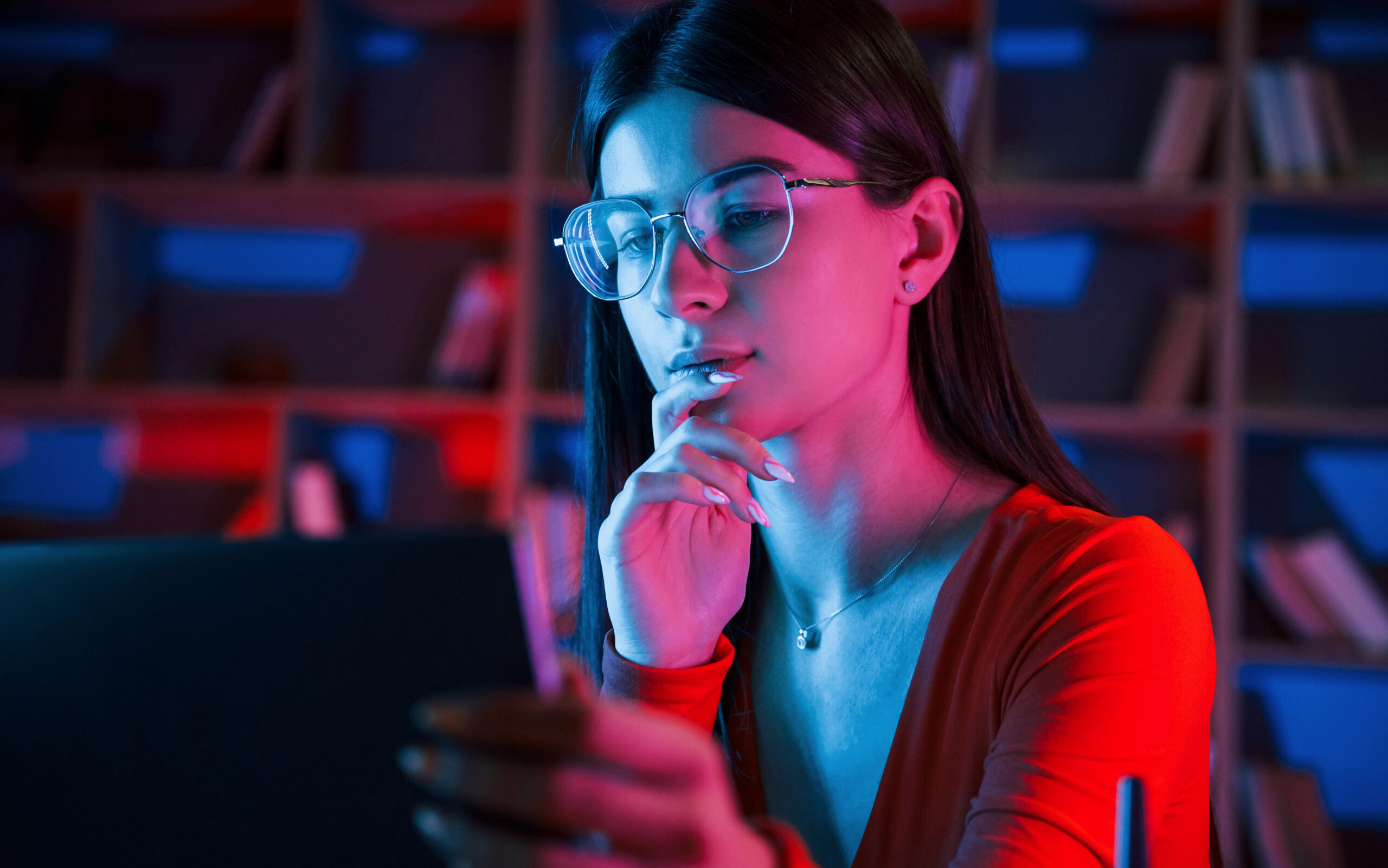 Beautiful pensive woman in glasses and red wear is sitting by the laptop in dark room with neon lighting