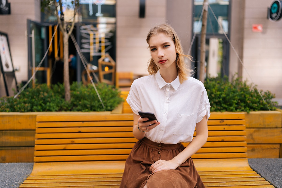 confident young woman sitting on bench holding mobile phone in hand in front of a building