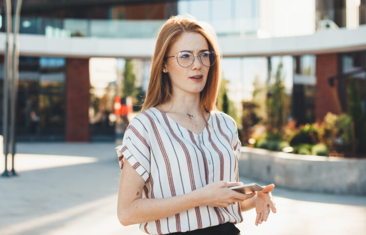 Confused woman with red hair and freckles wearing eyeglasses posing outside in the street holding a mobile and looking away