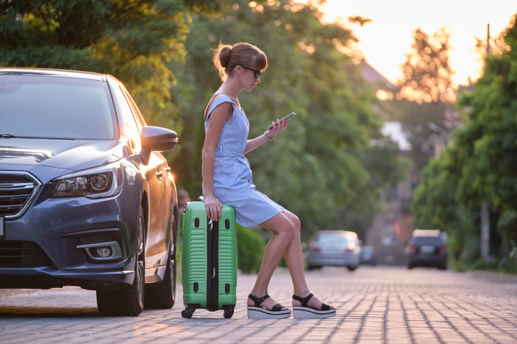 Young woman sitting on suitcase near her vehicle talking on her mobile phone on a city street in summer