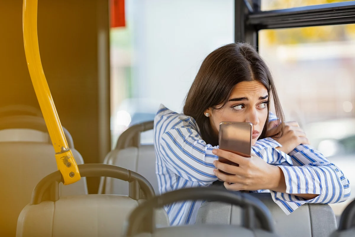 Young upset woman on a bus holding a cell phone looking out the window worried.