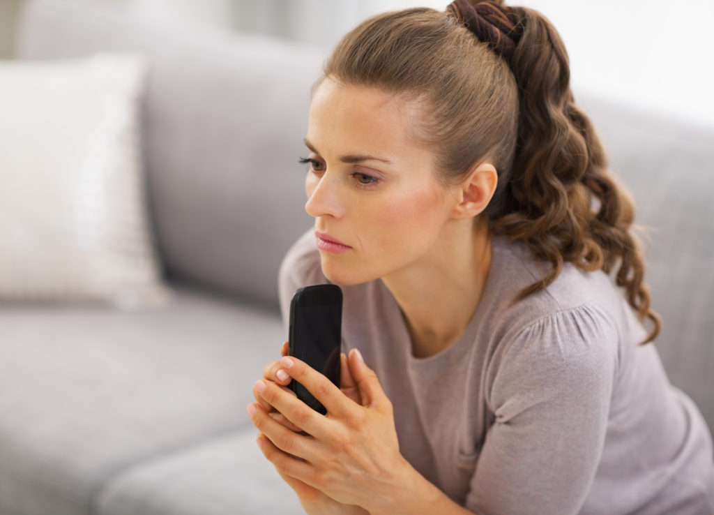 Concerned young woman sitting on sofa and holding mobile phone in hands