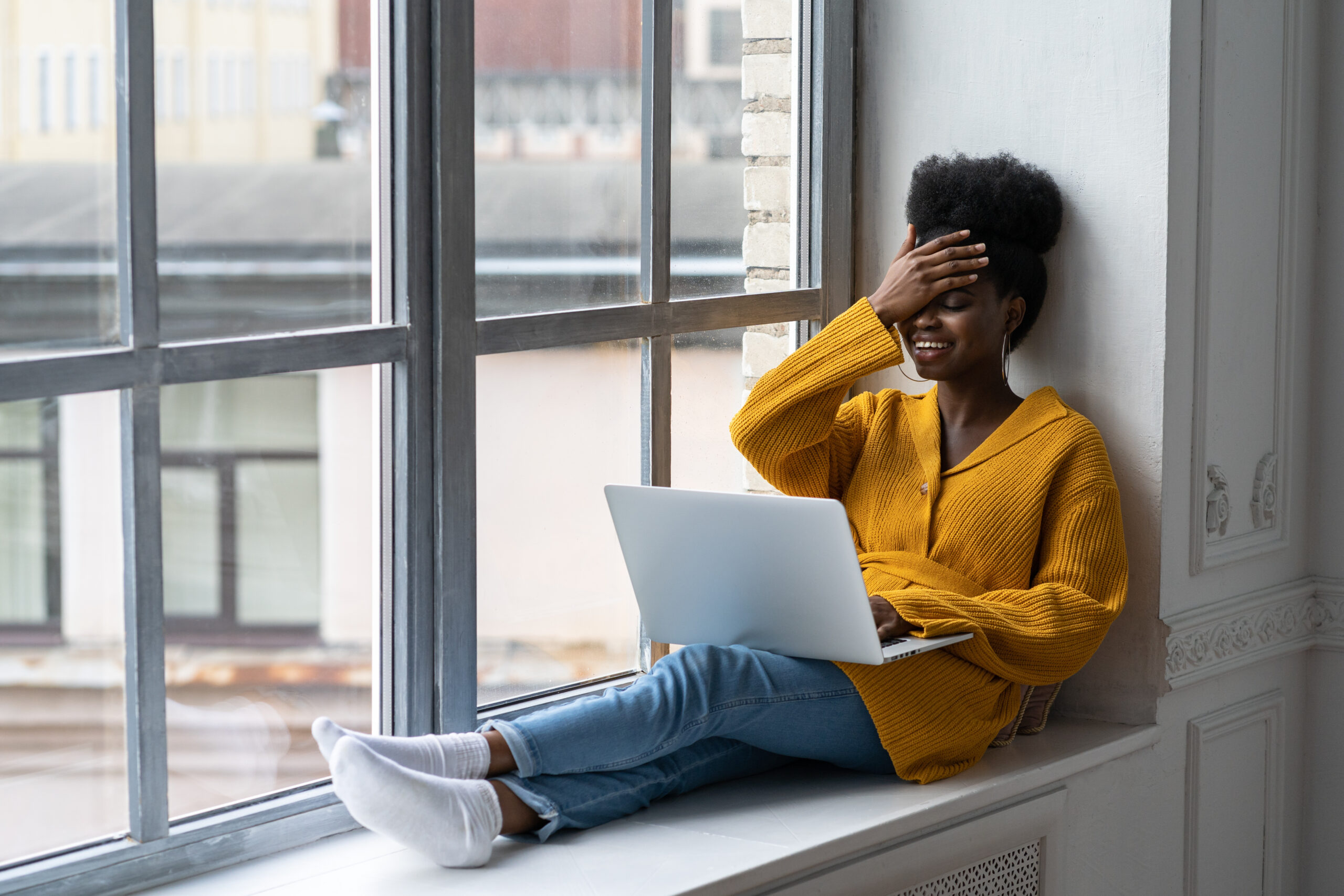 young woman sitting by the window facepalms while using laptop