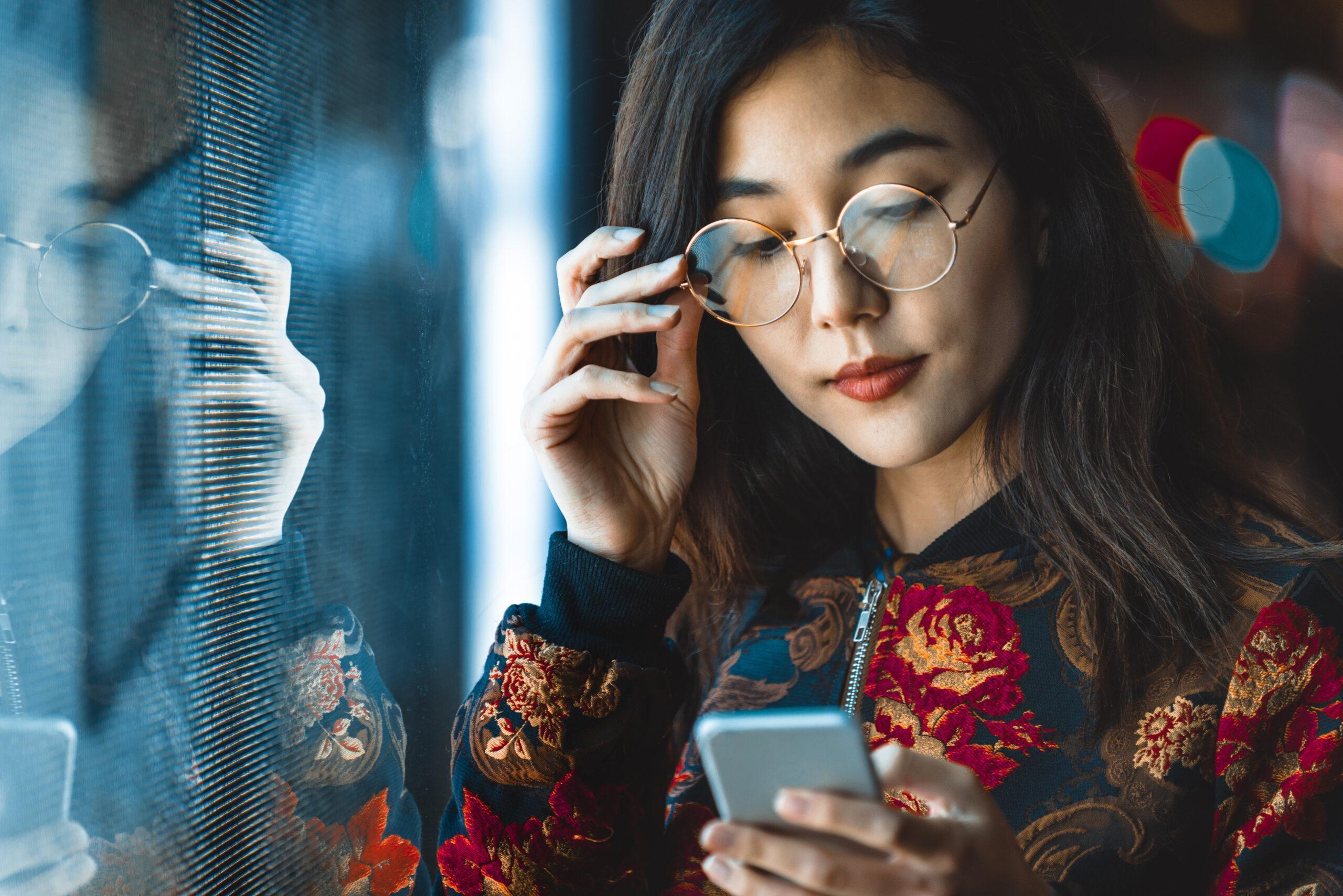 Pretty Asian woman  in a bright red blouse looking at her phone by a window