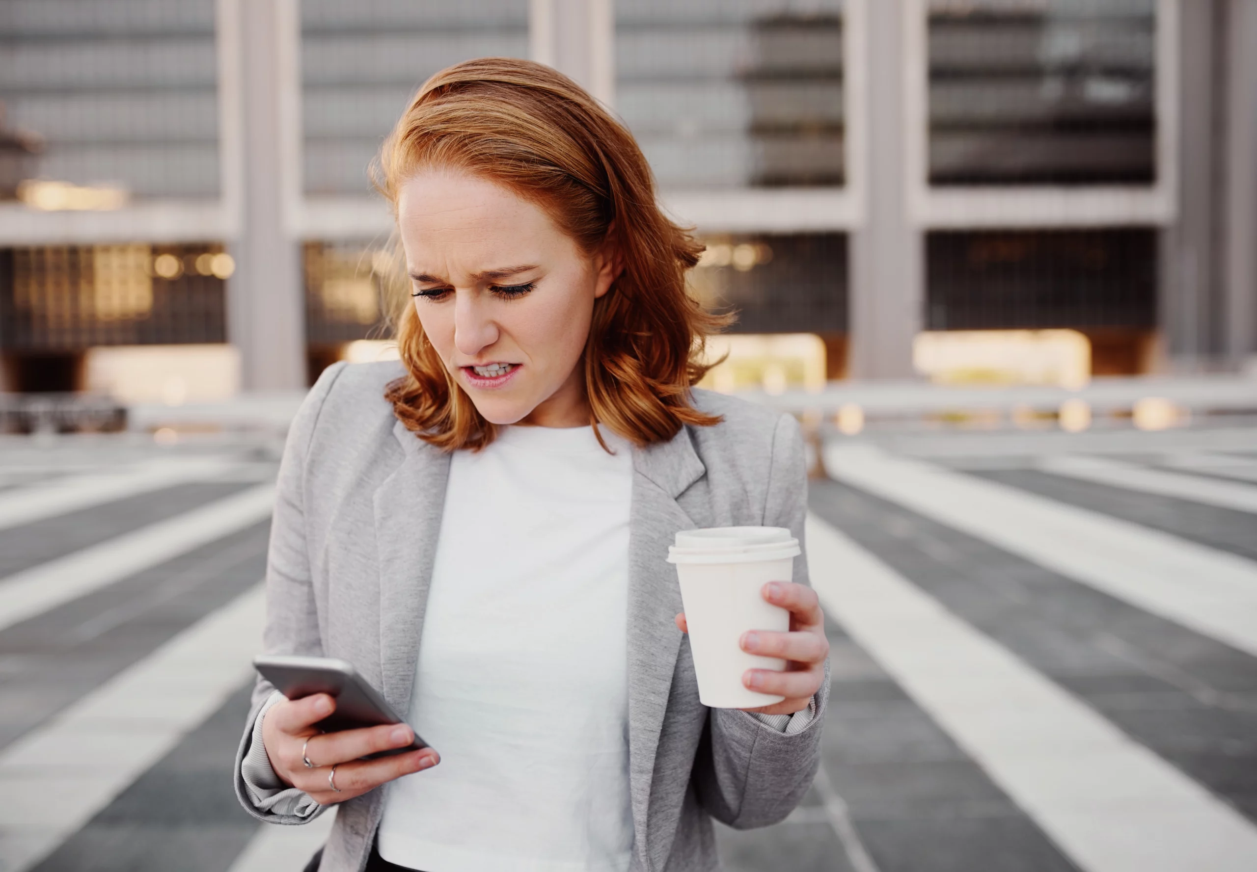 Frustrated young woman in formal clothing looking at smartphone while holding coffee cup