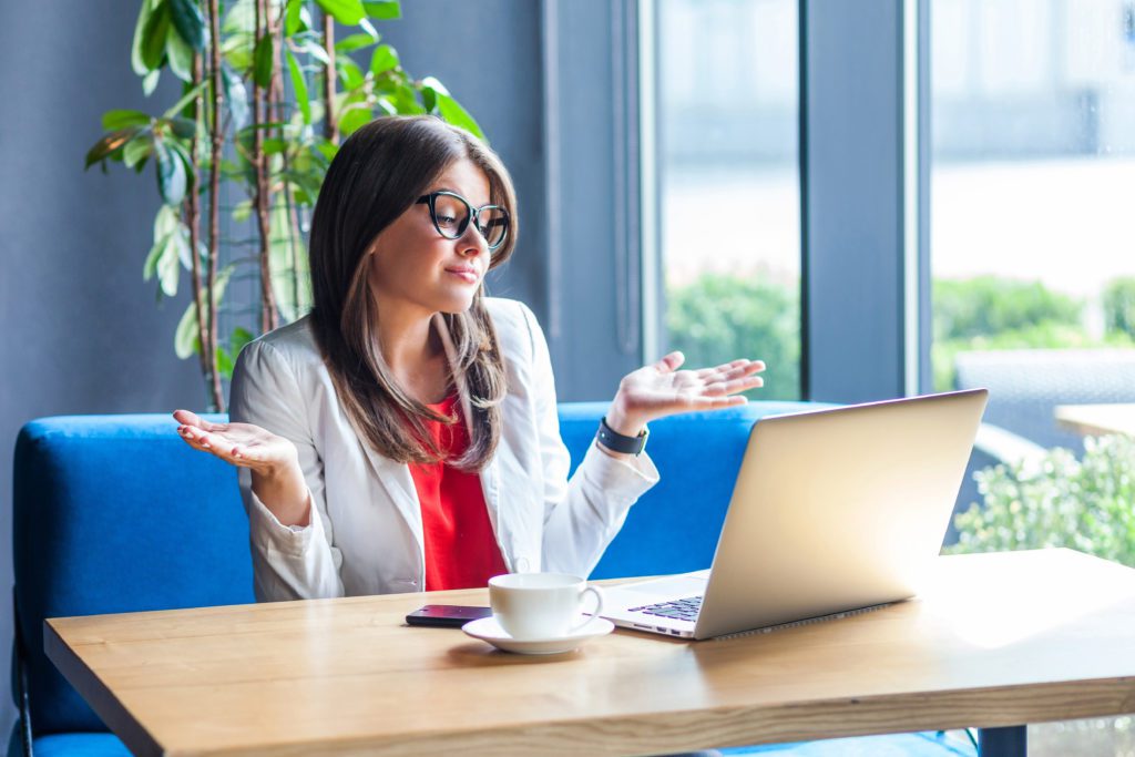 Woman doing a clueless gesture in front of her laptop, inside a cafe.