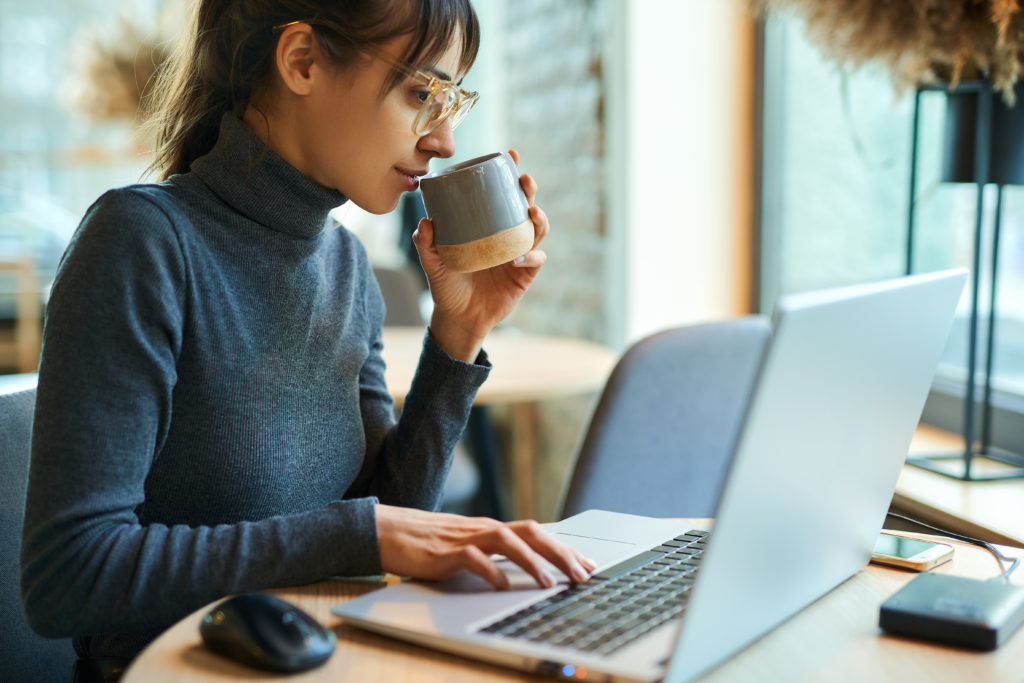 Young woman in eyeglasses enjoying coffee while working on portable laptop.