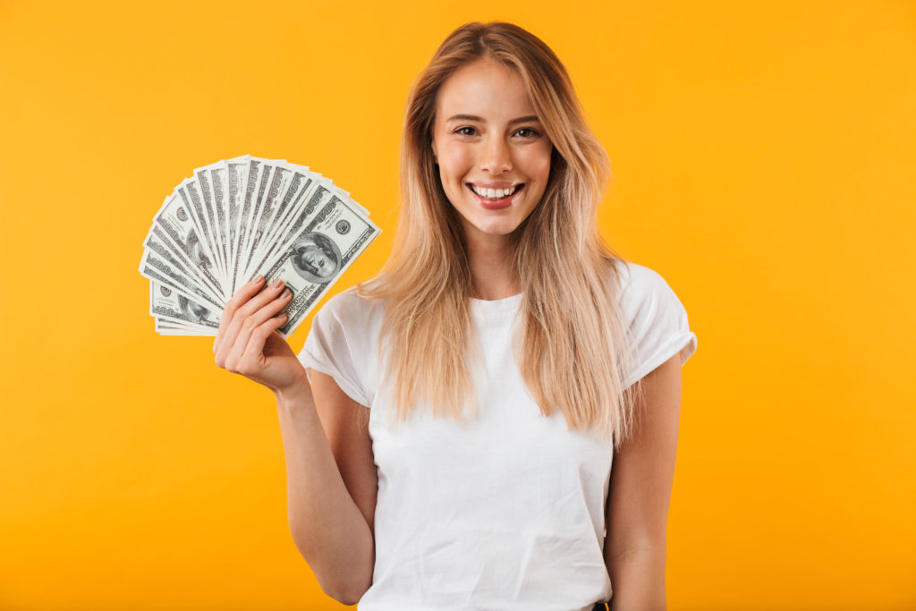 Portrait of a smiling young blonde girl showing bunch of money banknotes isolated over yellow background.