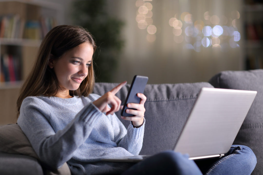 Woman smiling while using her phone and laptop at night.