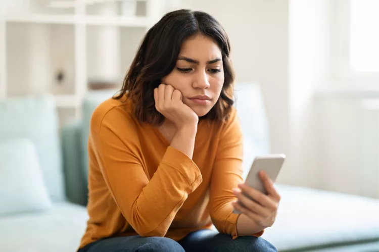 Pensive and upset young woman sitting on sofa looking intently at her cell phone