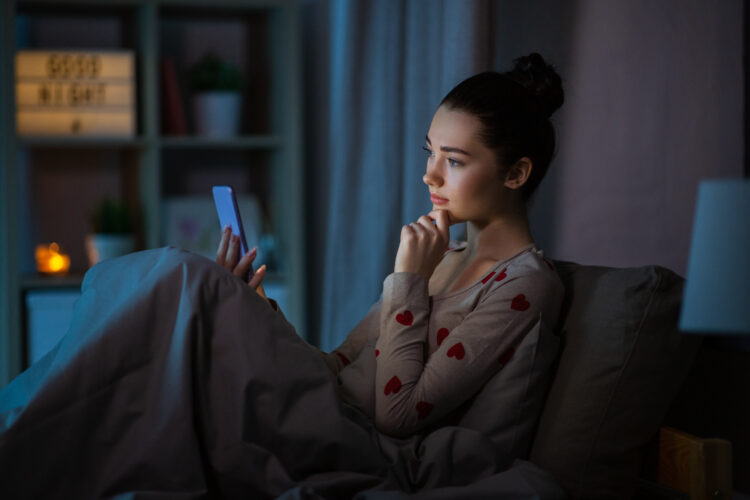 Young woman in pajamas with phone in bed at night, worried.