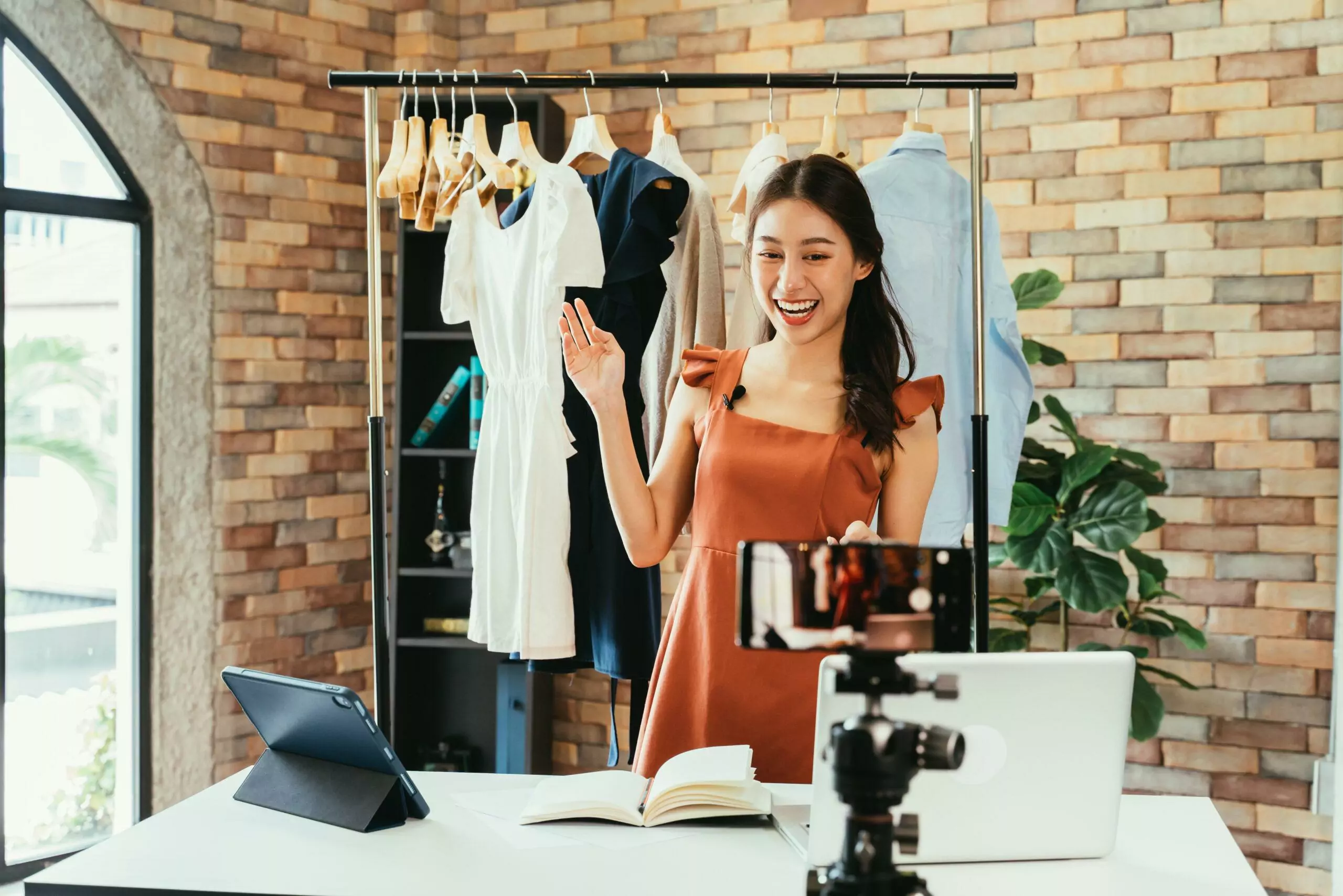 Young and beautiful Asian woman influencer showing clothes in front of smartphone camera while recording vlog video and live streaming at her shop