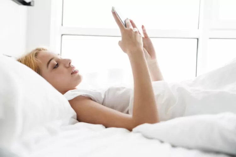 focused young woman using cellphone while lying in bed