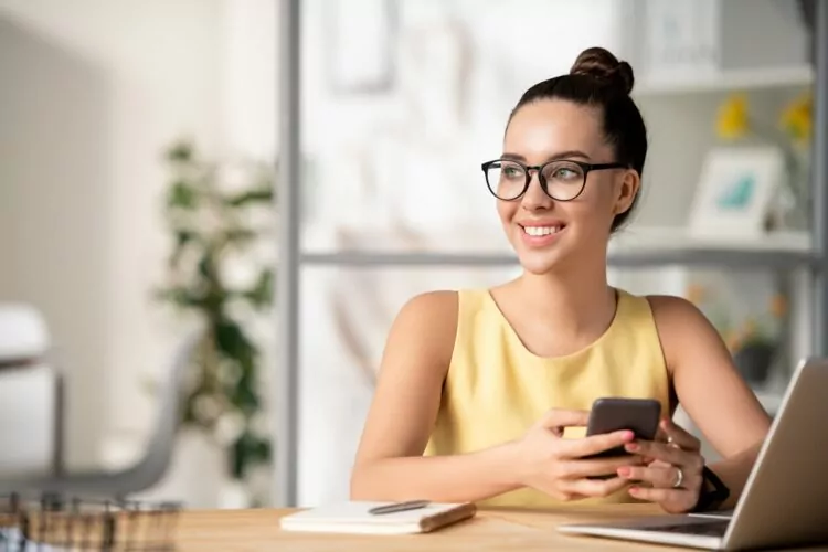 Smiling woman with glasses holding phone at home office trying to figure out how to turn of mock locations and get it working again.