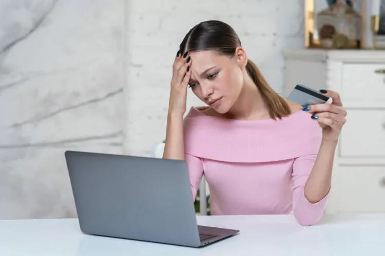 frustrated nervous woman is holding credit bank card in hand and looking at laptop computer.