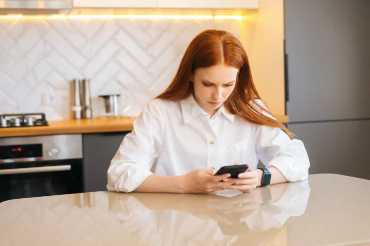 Serious attractive young woman using mobile phone sitting at table in kitchen.