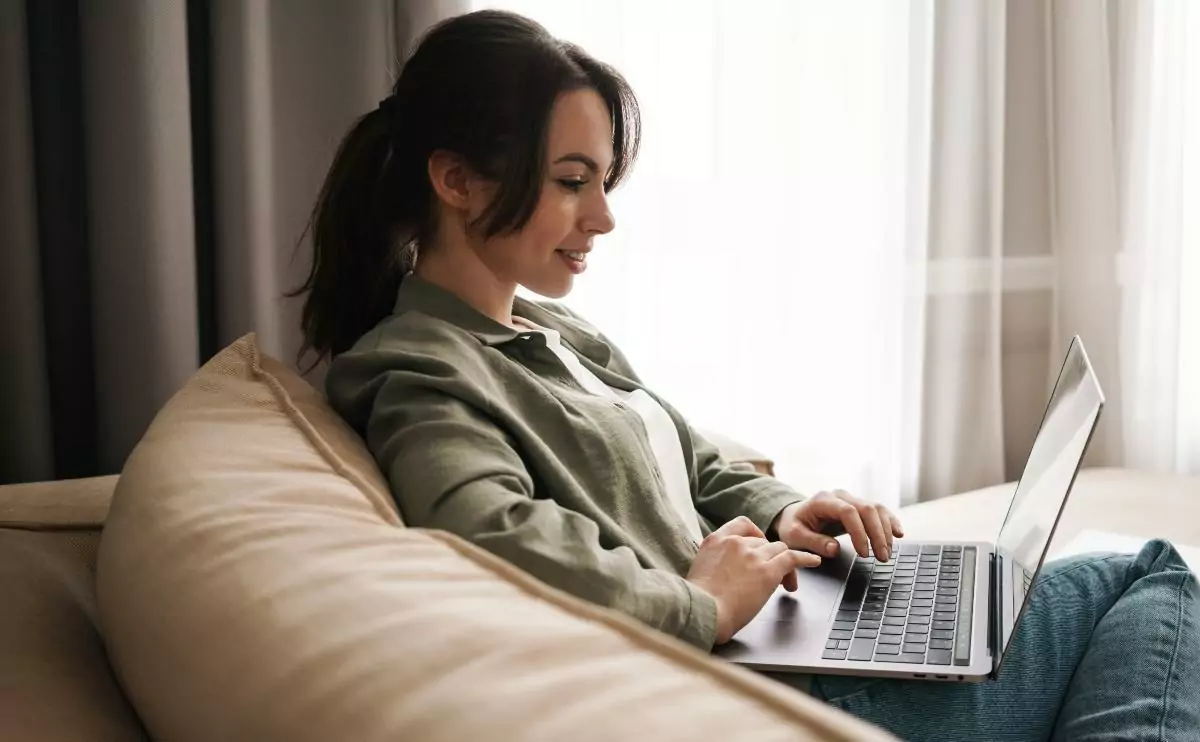 Brunette with hair tied in a ponytail sitting in a comfy couch and working on her laptop by the window