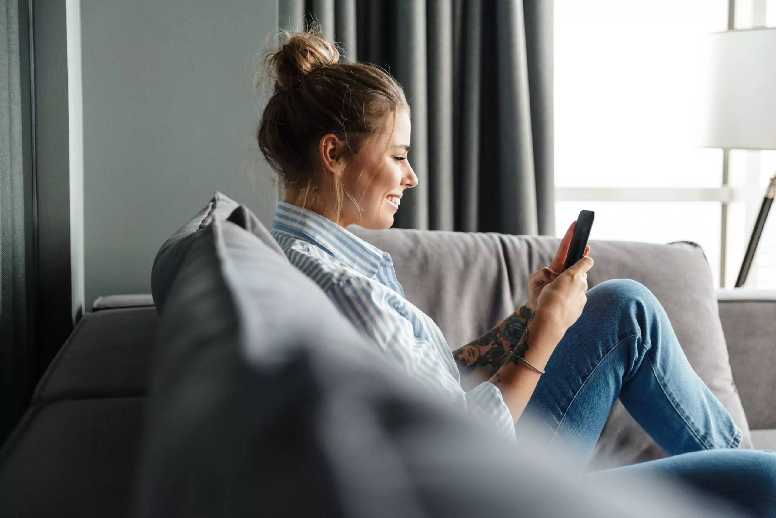 Image of happy young woman in striped shirt smiling and using cellphone while sitting on sofa at living room