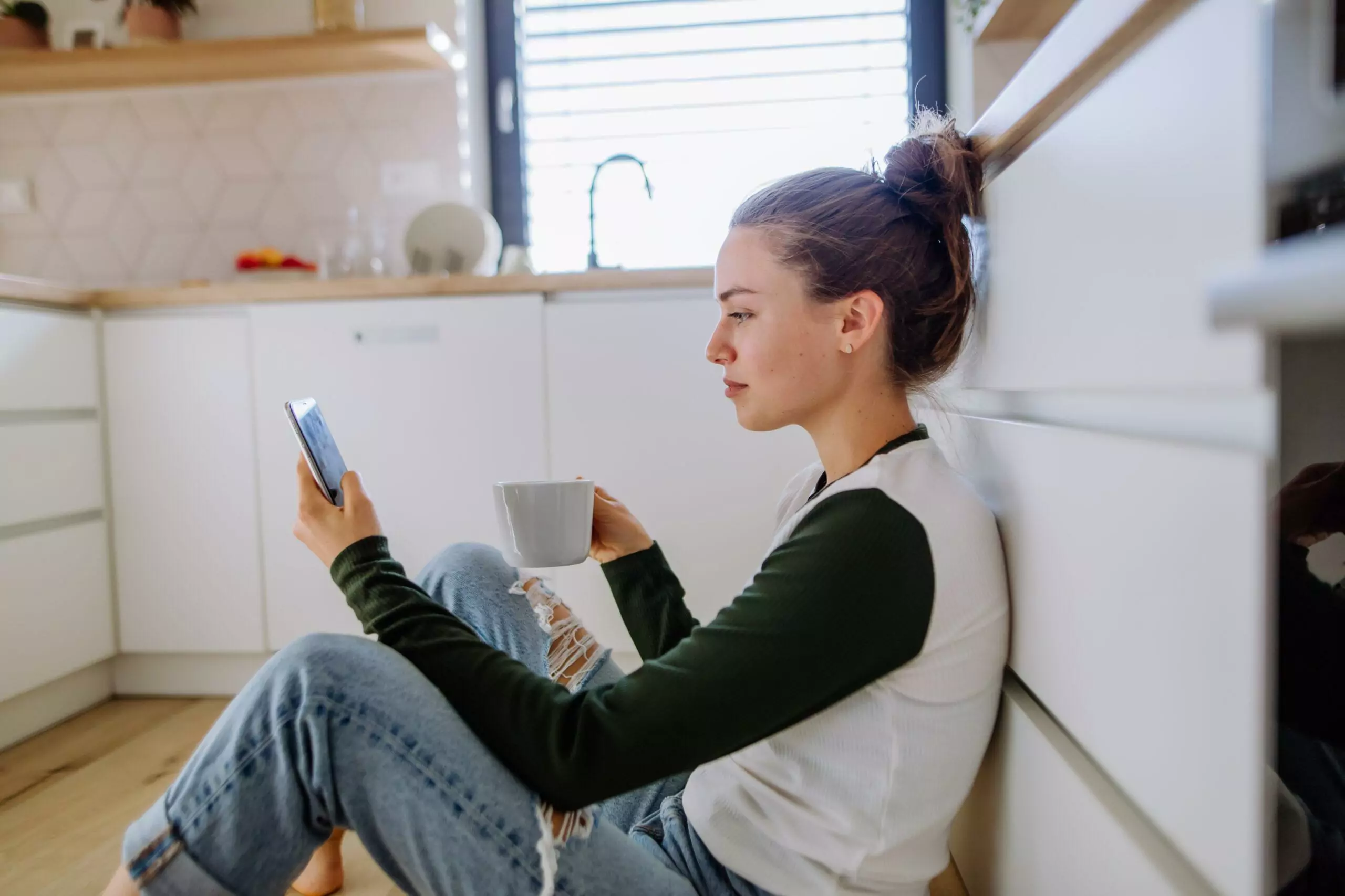 Young woman sitting with smartphone and cup of coffee in her kitchen