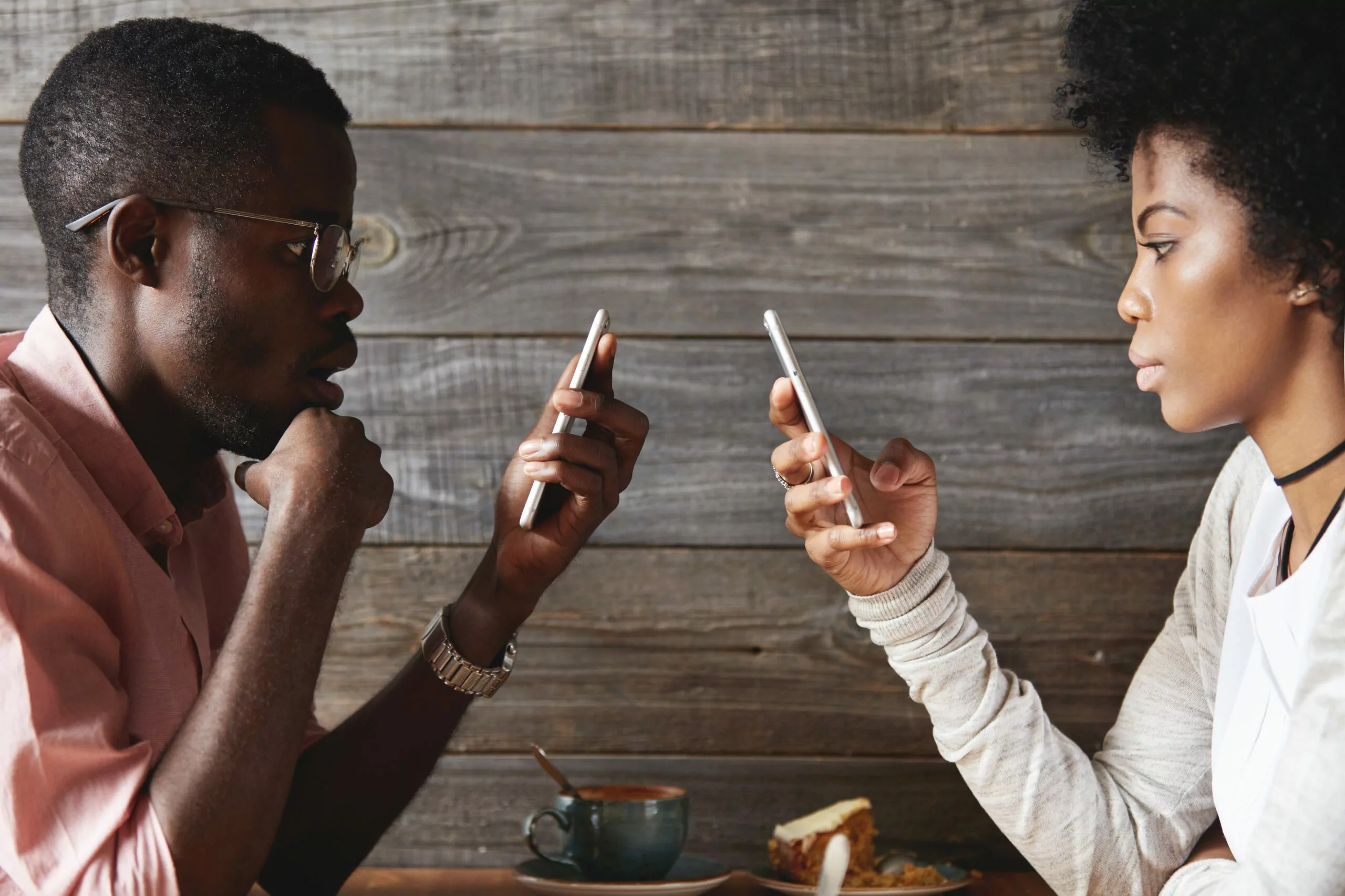 Internet addiction concept. Fashionable African couple using mobile phones totally absorbed in online life, looking obsessed, not talking to each other, using wi-fi at cafeteria, facing one another