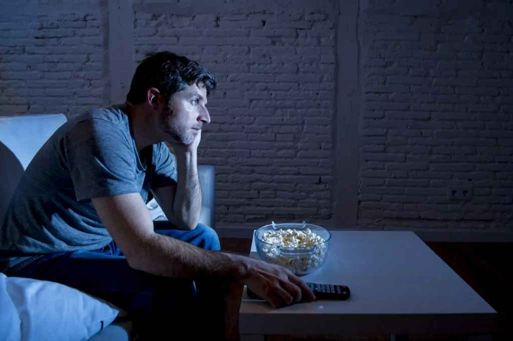 Man sitting on a sofa while watching television and eating popcorn at night.