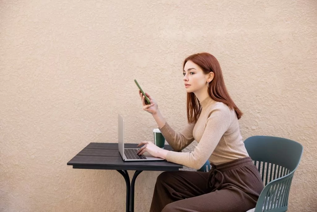 young redhead freelancer in turtleneck using smartphone near laptop on summer terrace.