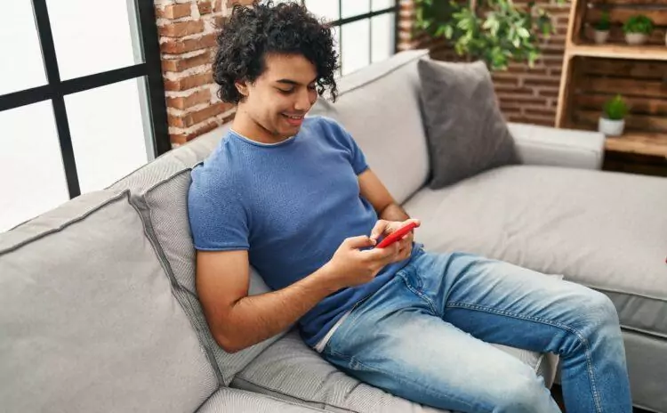 Young Hispanic man using smartphone sitting on sofa at home