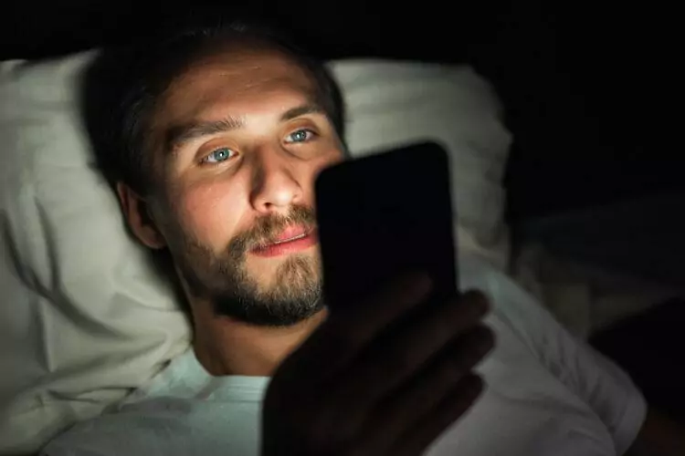 Young but tired man with a beard who cannot sleep and is watching a video on his smartphone
