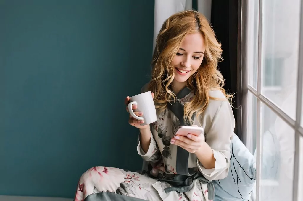 Pretty blonde girl sitting on window sill with cup of coffee, tea and smartphone in hands.