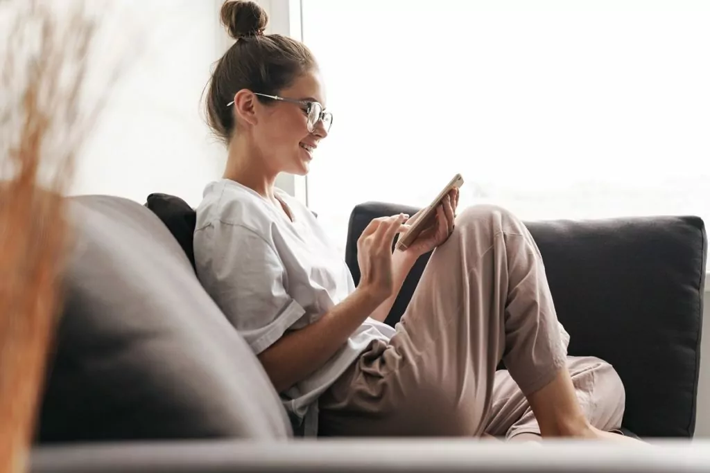 Beautiful woman smiling and typing on cellphone on sofa.