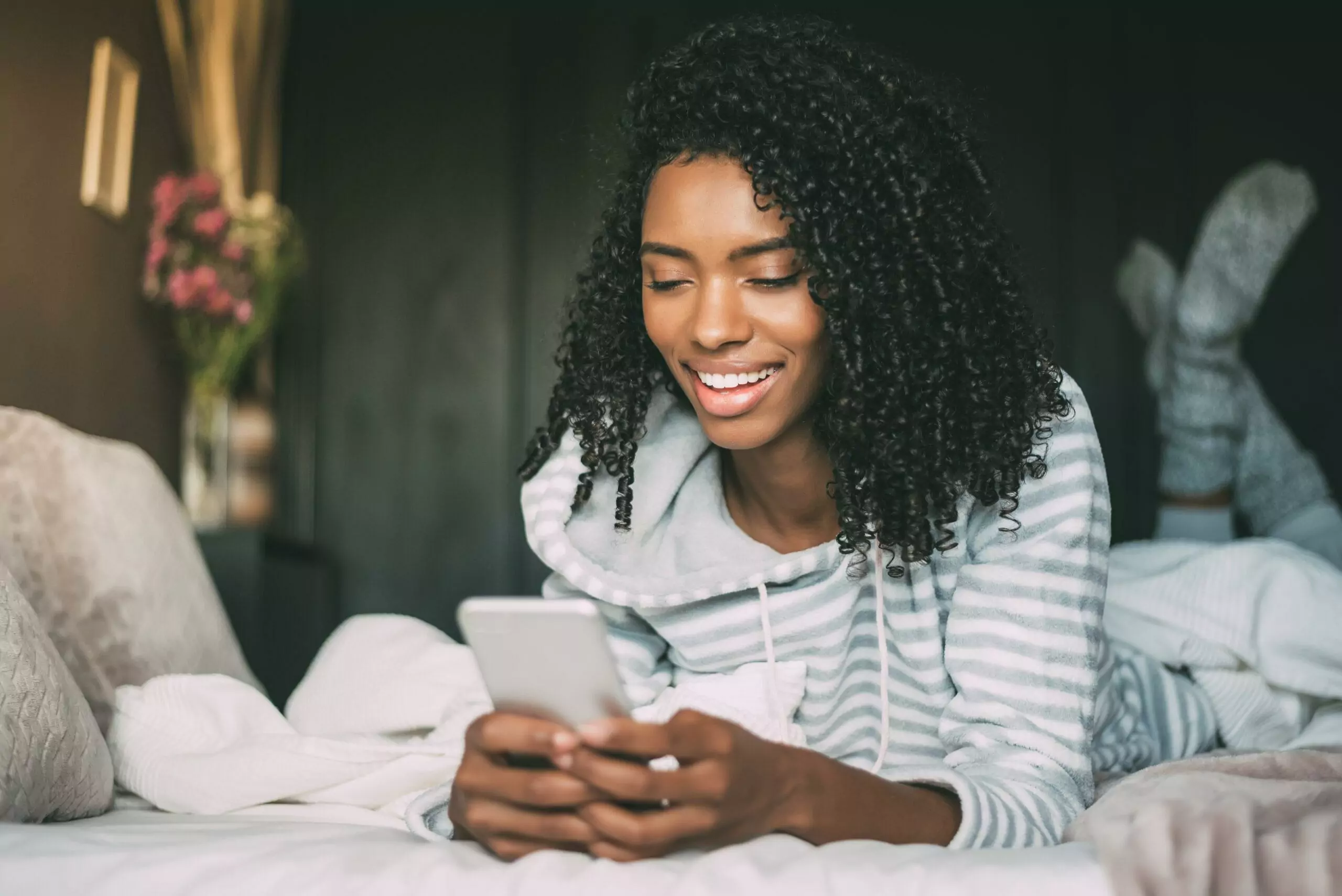 Pretty African-American woman with curly hair smiling and using her smartphone in bed