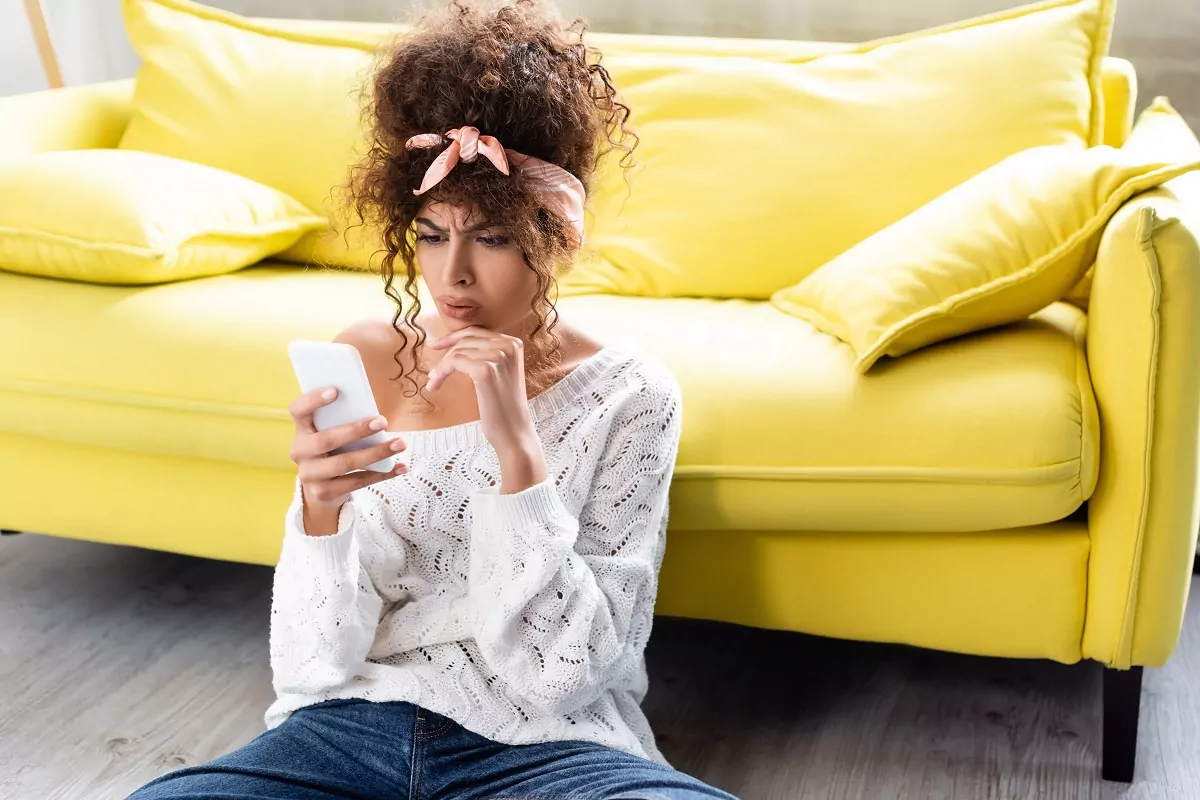 worried woman looking at smartphone near yellow sofa