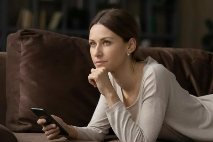Young serious woman looking pensive lying on sofa with smartphone