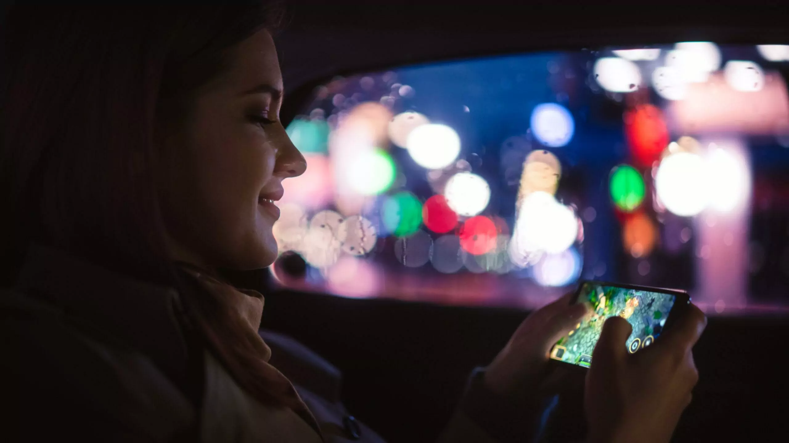 Happy female is playing with her smart phone while commuting home in a backseat of a taxi at night