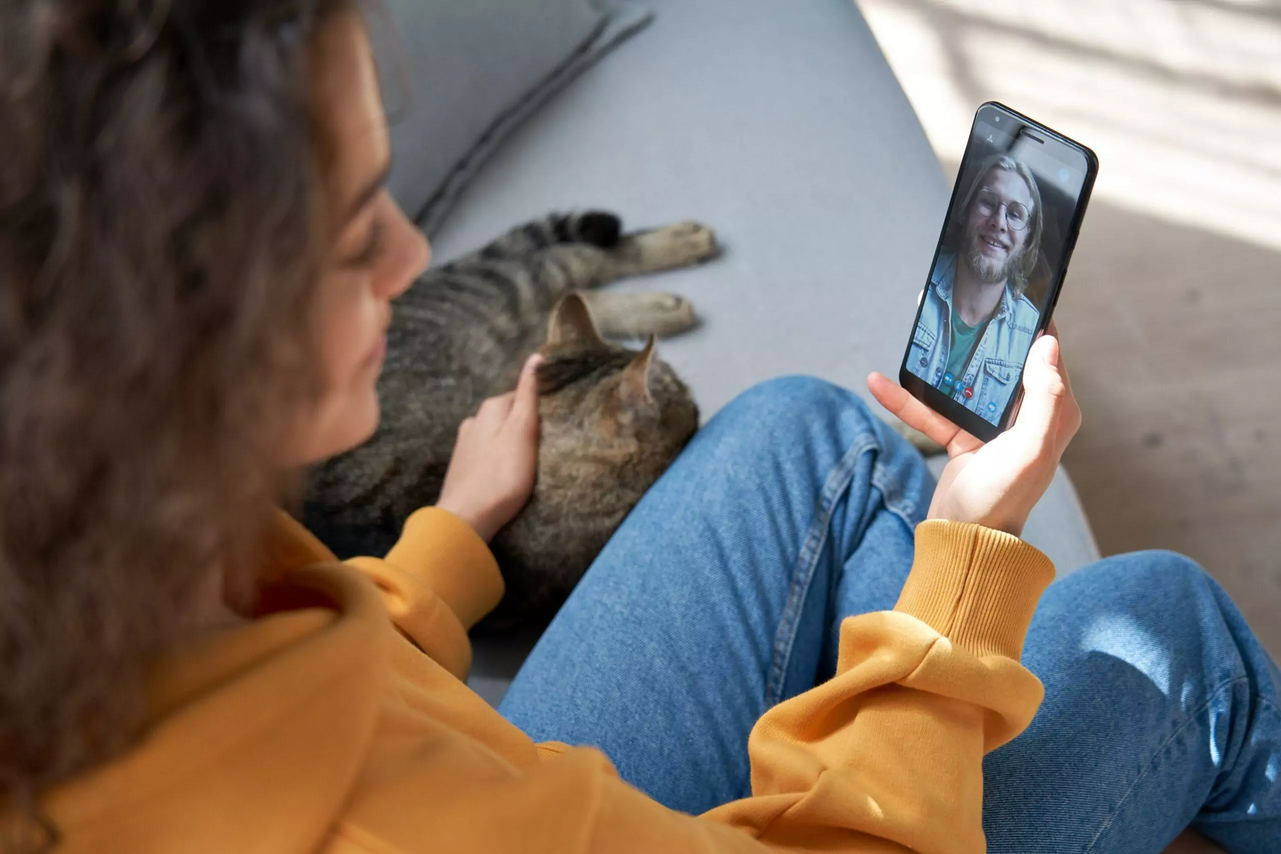 Young hispanic girl sit on sofa with cat hold phone video call on mobile screen.
