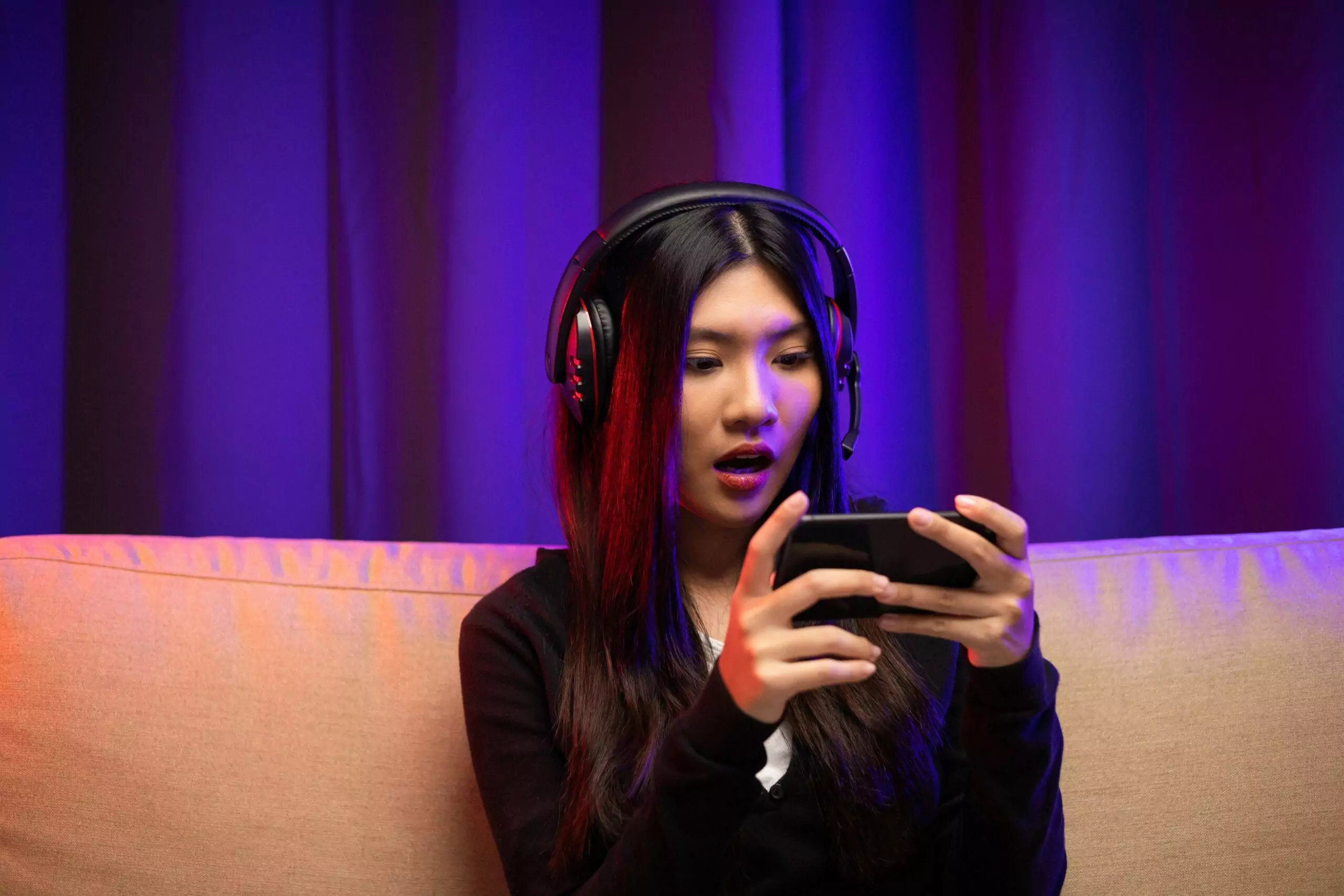 Shocked Asian female gamer with headphone holding smartphone and playing games 