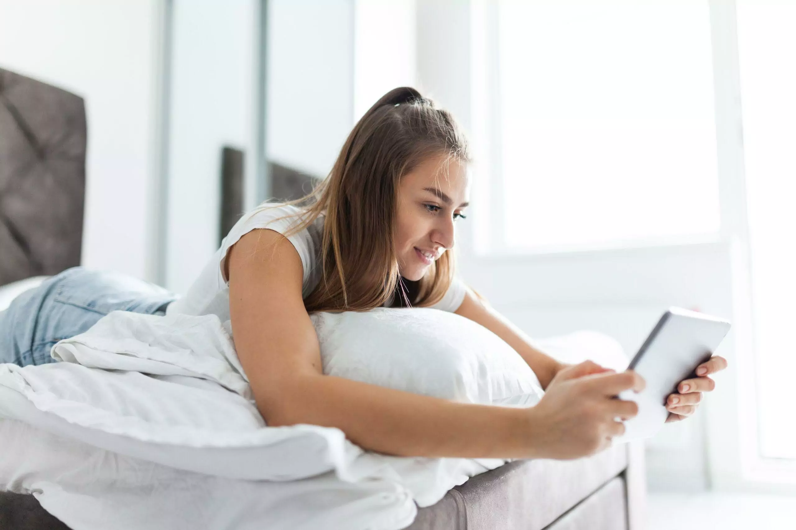 Young beautiful caucasian woman uses a tablet while lying on a bed in a bright room